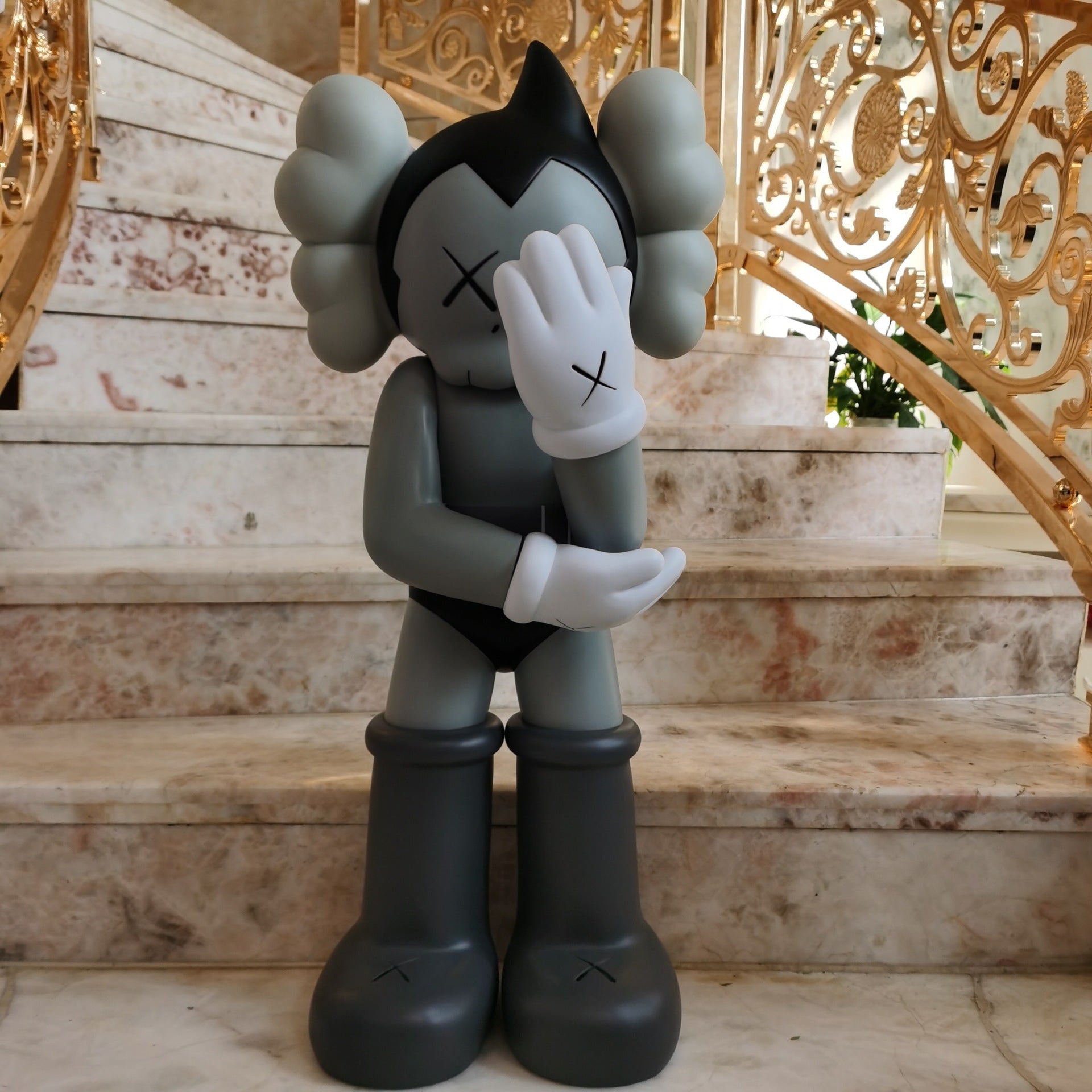 A gray and black cartoon-style collectible figure with white gloves and black boots, standing on marble stairs in front of an ornate gold railing.