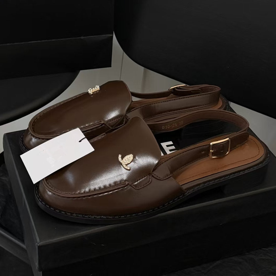 A pair of dark brown leather slingback loafers with gold-tone hardware and a small metallic crest emblem on the toe, resting on a black shoebox.