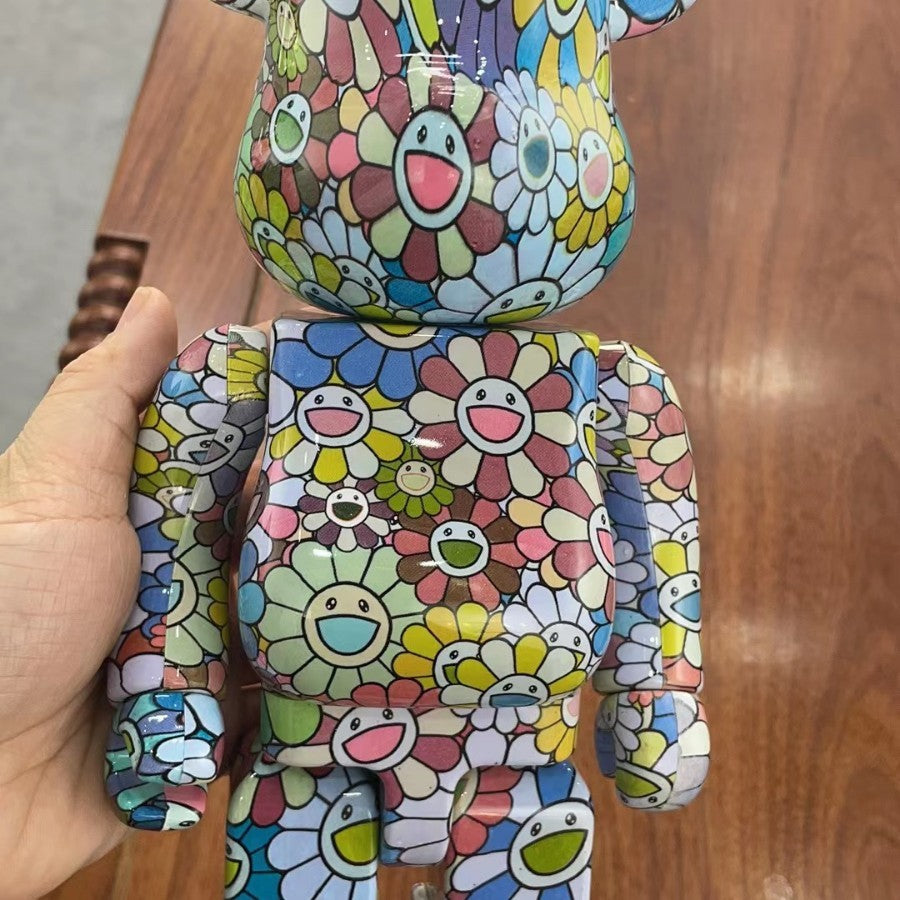 A hand holding a large bear-shaped collectible figure covered in a colorful floral mosaic pattern, photographed over a wooden tabletop.