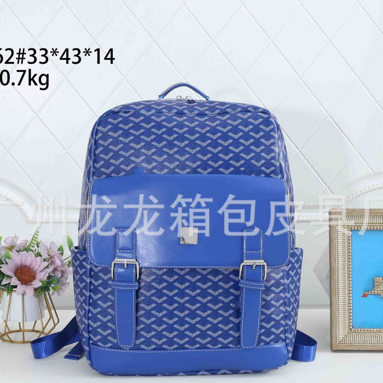 A blue patterned backpack with a glossy front flap and buckle straps, displayed on a white surface with decorative flowers and a picture frame in the background. Text overlays show product details and a website watermark.