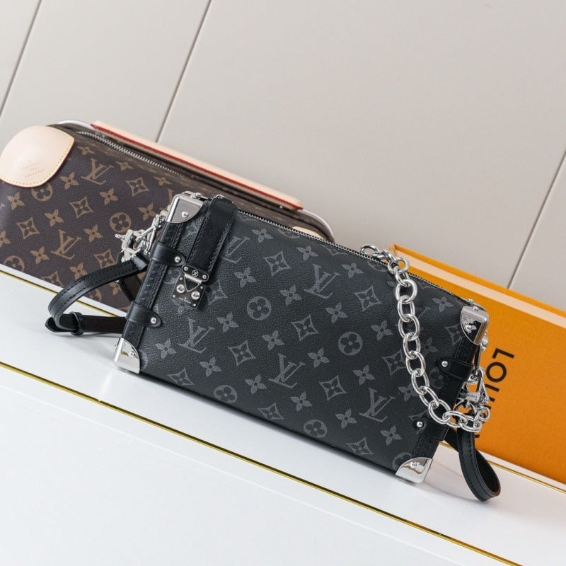 A Louis Vuitton shoulder bag in black monogram canvas with silver-tone chain and leather trim, displayed on a white surface with a second bag and an orange box in the background.