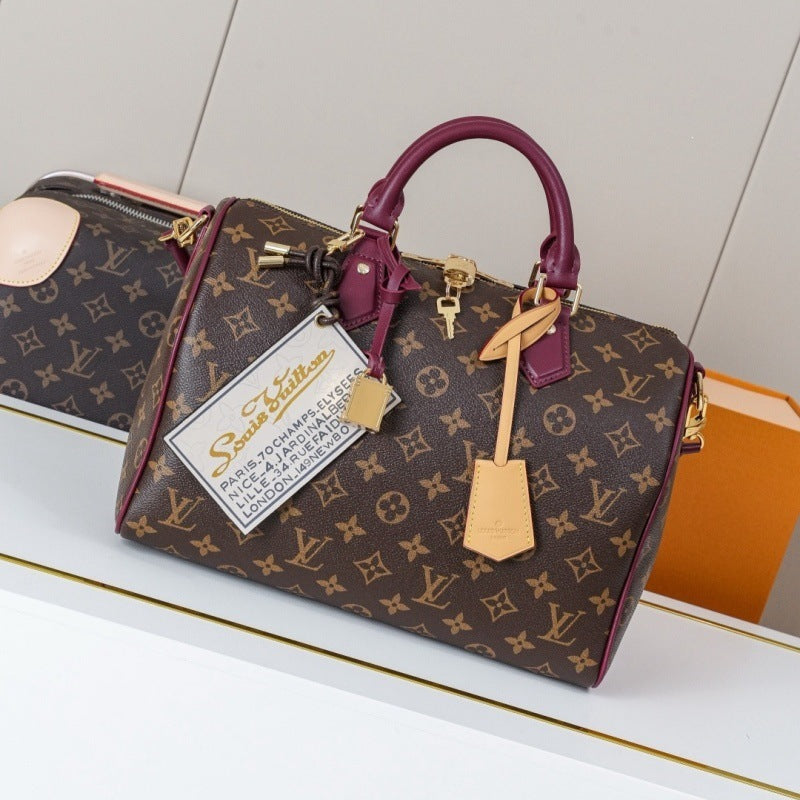 A Louis Vuitton Speedy bag in brown monogram canvas with dark magenta leather trim and handles, displayed on a white surface with a matching bag and orange box in the background.