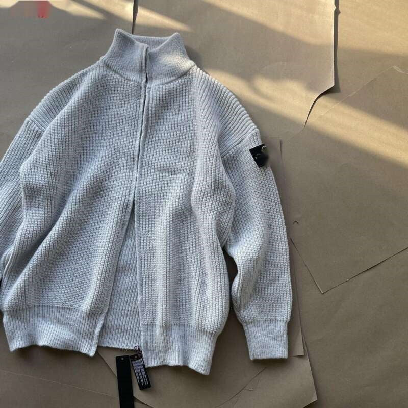 A light gray ribbed knit zip-up jacket with a mock neck collar, laid flat on brown paper packaging. A black woven brand patch is visible on the left sleeve, and a hangtag is attached near the hem.