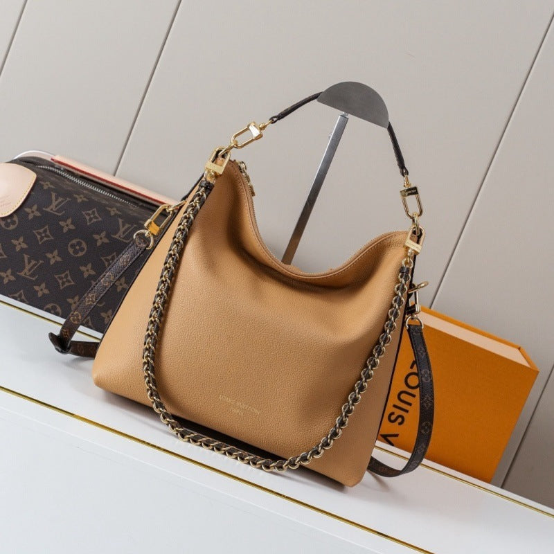 A tan pebbled leather shoulder bag with a gold-tone chain strap and black leather handle, displayed on a white shelf next to a Louis Vuitton monogram zip case and an orange Louis Vuitton box.
