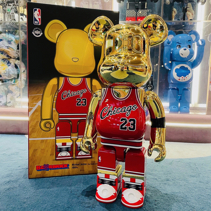 A shiny gold Bearbrick toy wearing a red Chicago Bulls jersey with the number 23 stands in front of its original packaging box. A blue Care Bear figure is visible in the background inside a display case.