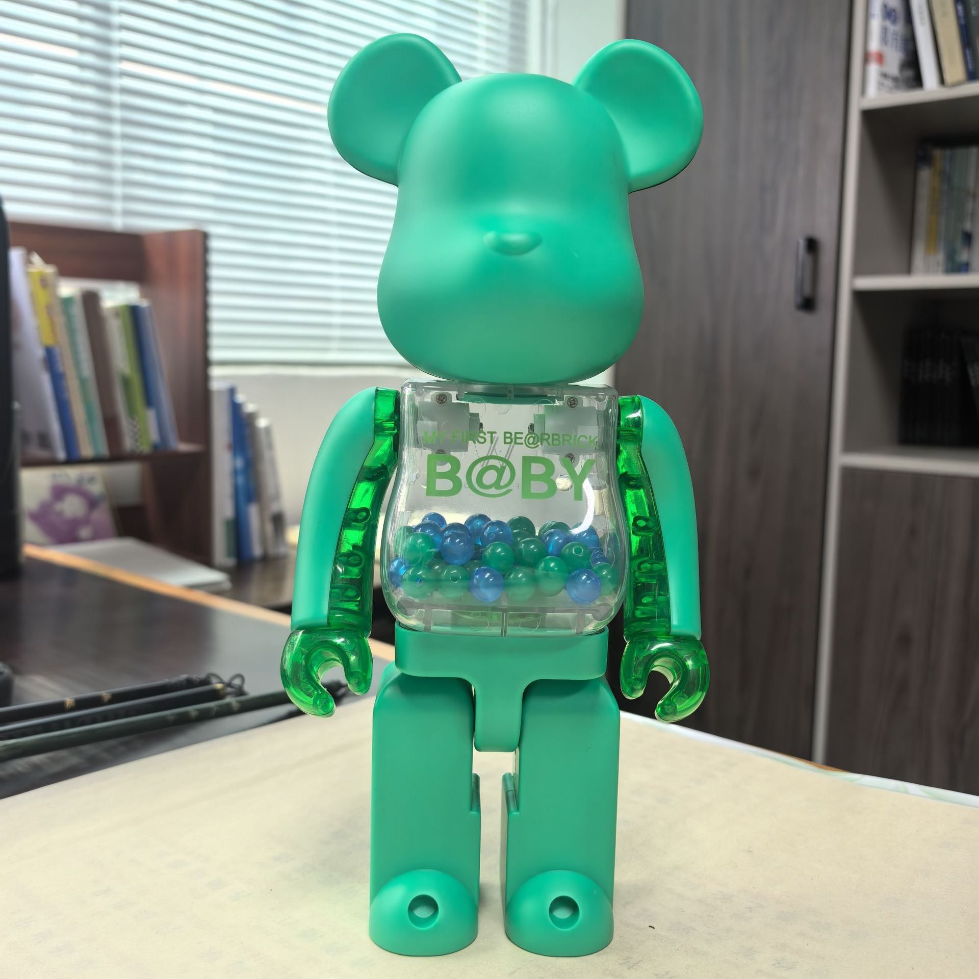 A translucent mint green bear-shaped toy figure with a clear torso filled with blue and green beads, standing on a tabletop in an indoor room with bookshelves in the background.