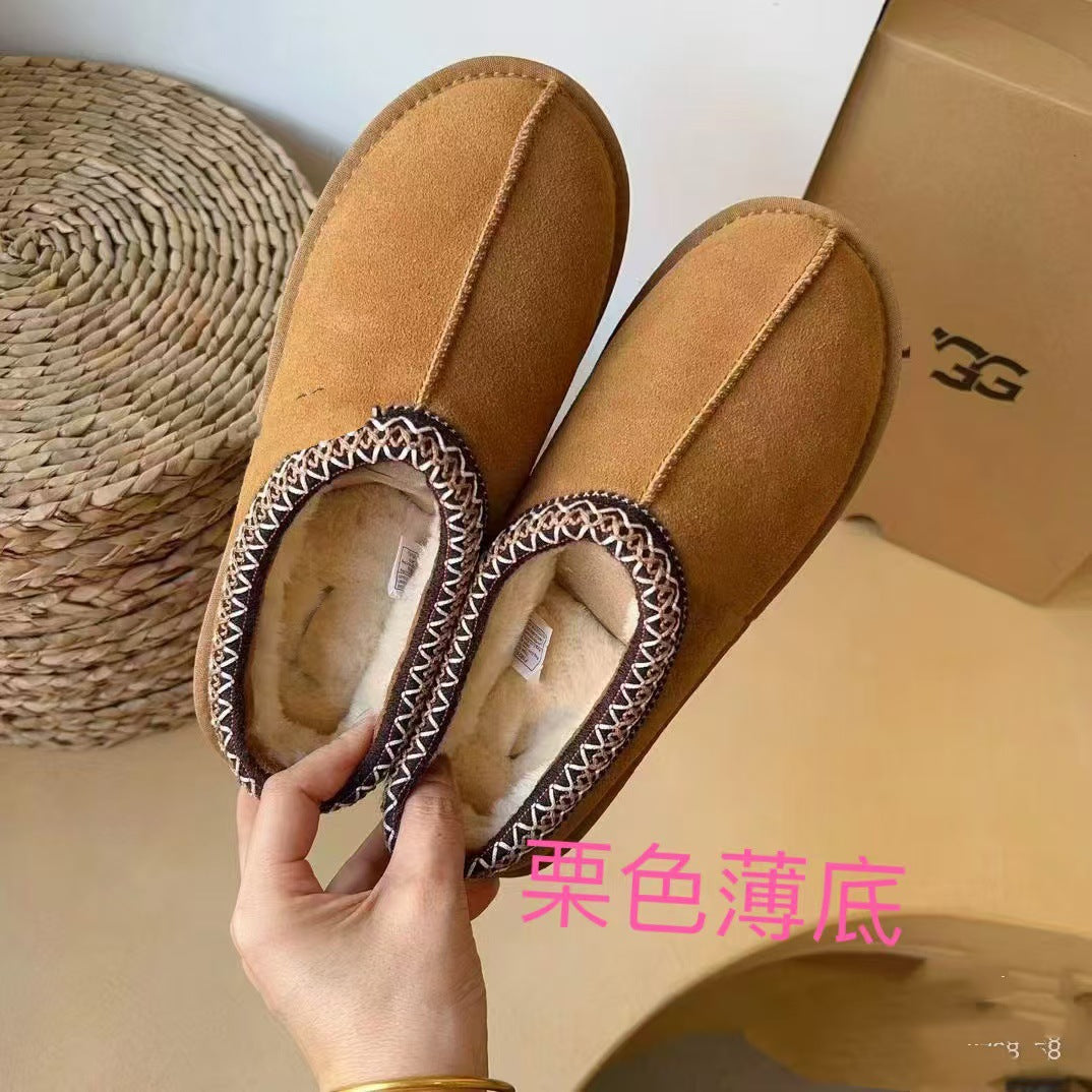 A pair of tan suede slip-on slippers with a white and black zigzag patterned trim, held by a hand. A woven mat is on the left, and a cardboard box is on the right.