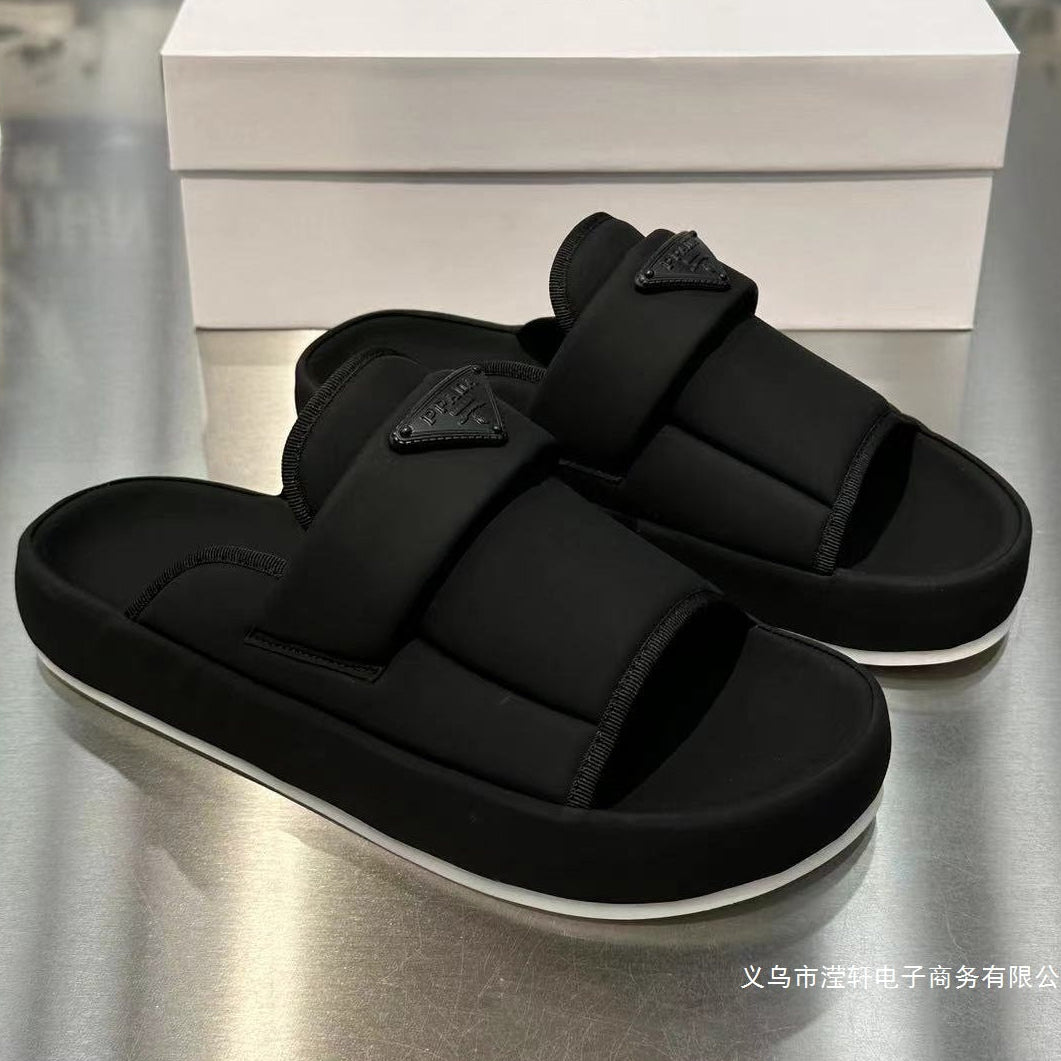 A pair of black slide sandals with wide padded straps and a small triangular logo plaque, displayed on a metallic surface with a white shoe box behind them. The box shows the brand name 'Zhu Zhaolin'.