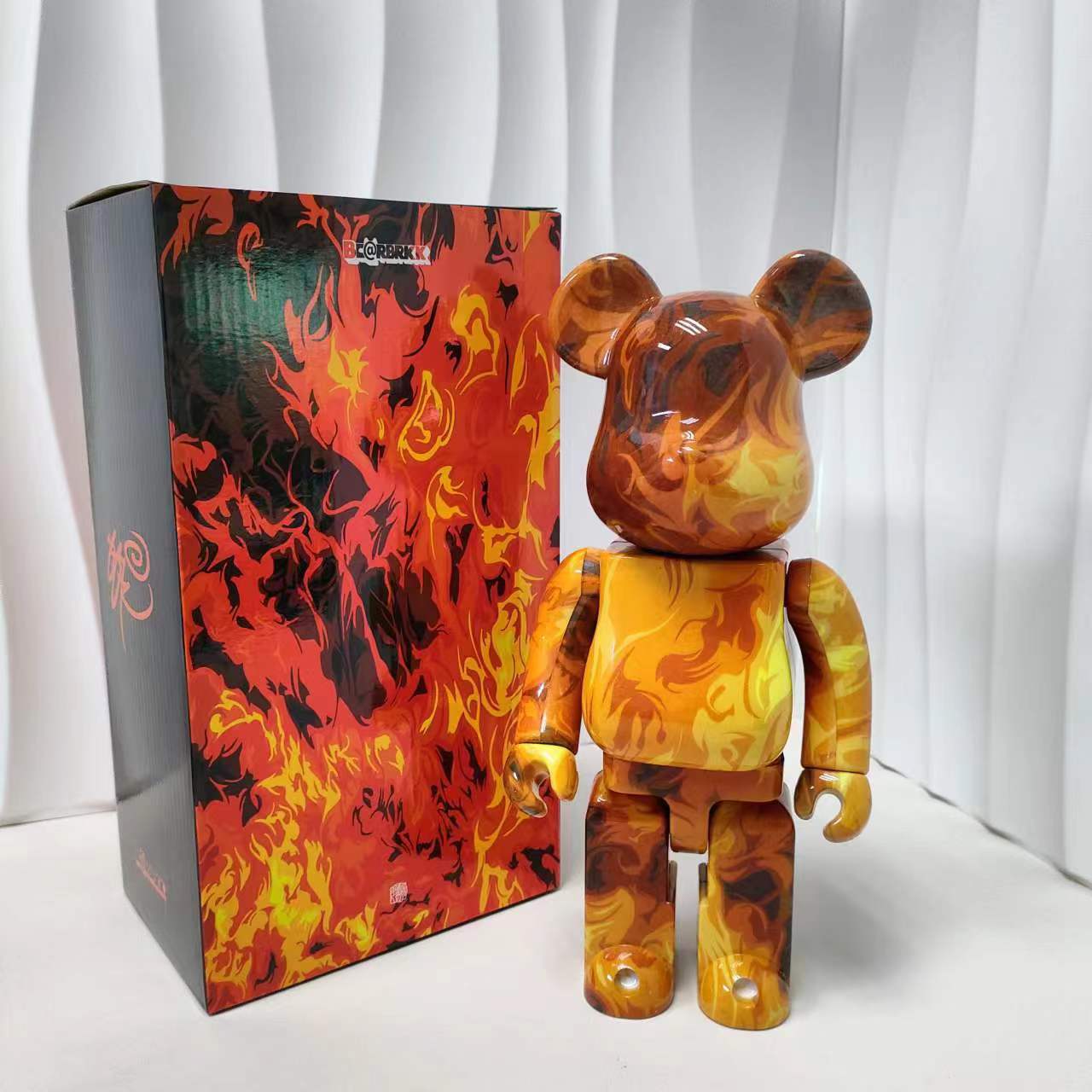 A collectible bear figure with a glossy orange, yellow, and brown flame marbled paint job stands next to its matching retail box, which features a matching fiery pattern, set against a white curtain background.