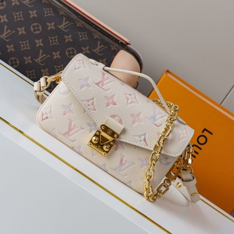 A small Louis Vuitton shoulder bag in off-white monogram canvas with gold-tone hardware, displayed on a white surface next to an orange Louis Vuitton box and a dark monogram pouch.