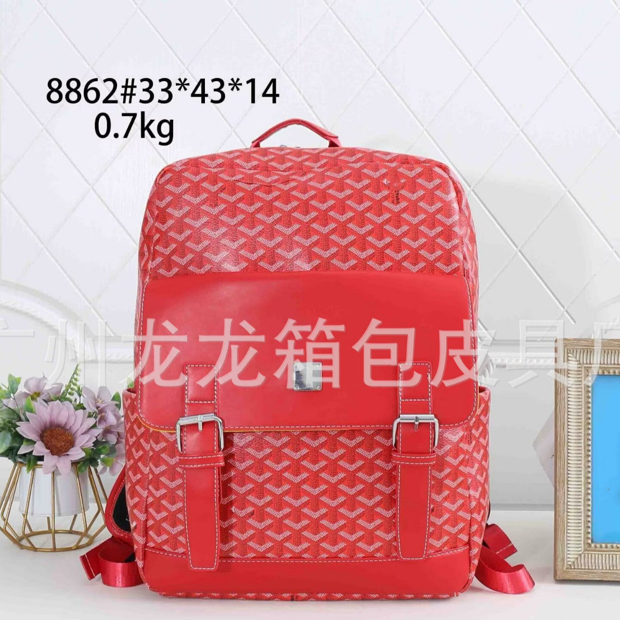 A red faux leather backpack with a repeating geometric monogram pattern, featuring a front flap with two silver buckles and red ribbon details. The image is set against a white tiled wall background with decorative props, including a small flower arrangement and a picture frame.
