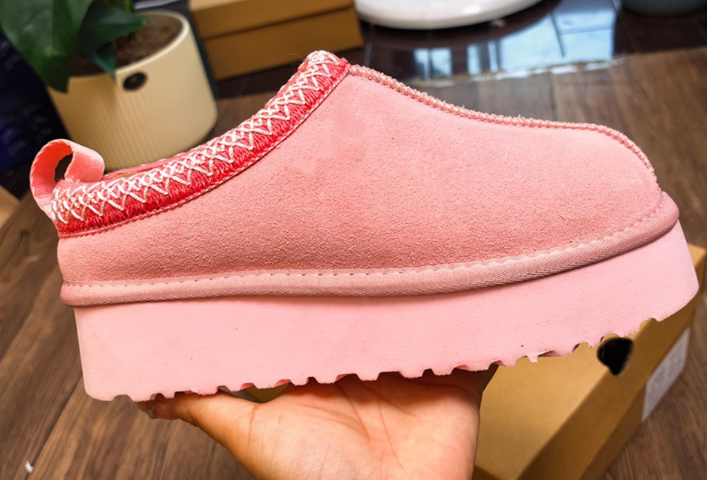 A pink suede slip-on clog shoe with a red and white patterned trim and a chunky platform sole, held in a hand over a wooden floor background.