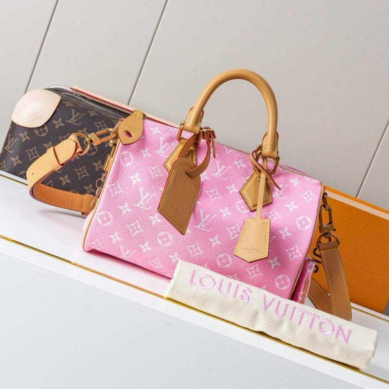 A pink Louis Vuitton monogram Speedy handbag with tan leather handles and trim, displayed on a white shelf alongside a brown Louis Vuitton monogram bag and an orange box, with a white dust bag reading 'LOUIS VUITTON' in pink in front.
