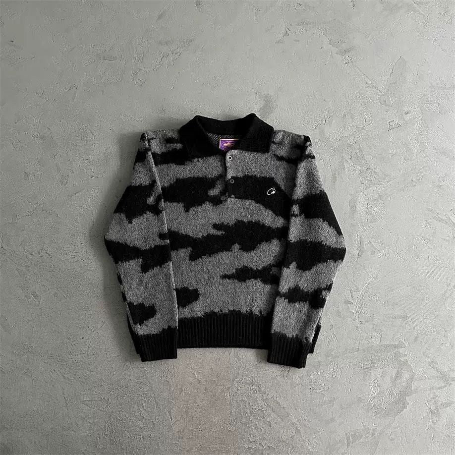 A black and gray patterned knit polo sweater with a black collar, ribbed cuffs, and a small white embroidered logo on the chest, laid flat on a textured gray background.