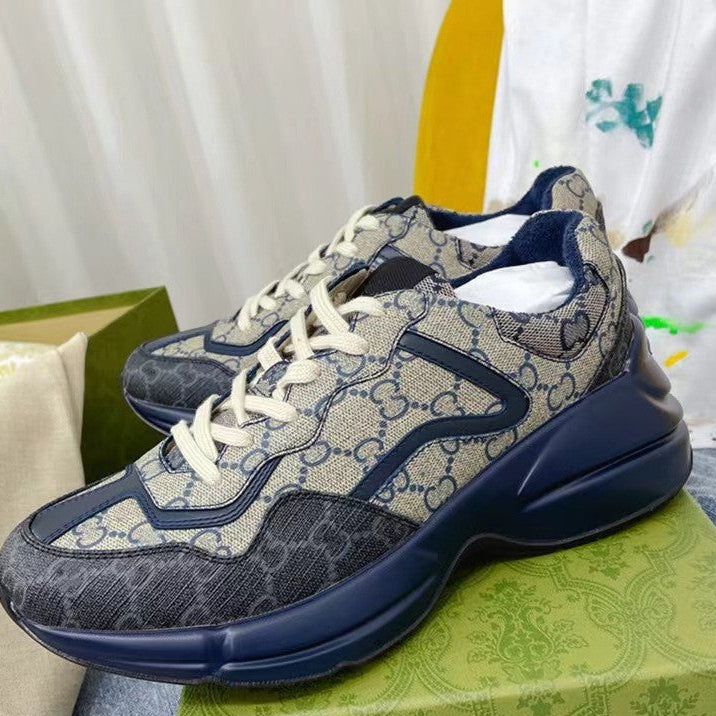 A pair of Gucci low-top sneakers with the brand's signature GG monogram pattern in beige and navy blue, featuring thick navy soles and cream-colored laces. The shoes are displayed on top of a green patterned shoebox.