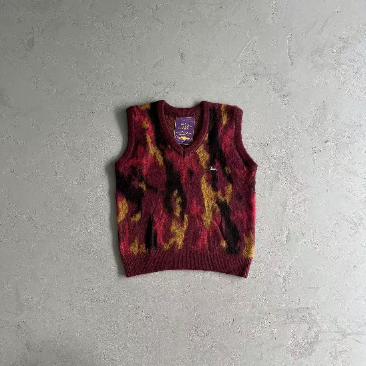 A sleeveless knit sweater vest with a maroon, yellow, and black tie-dye pattern, laid flat on a textured gray background.