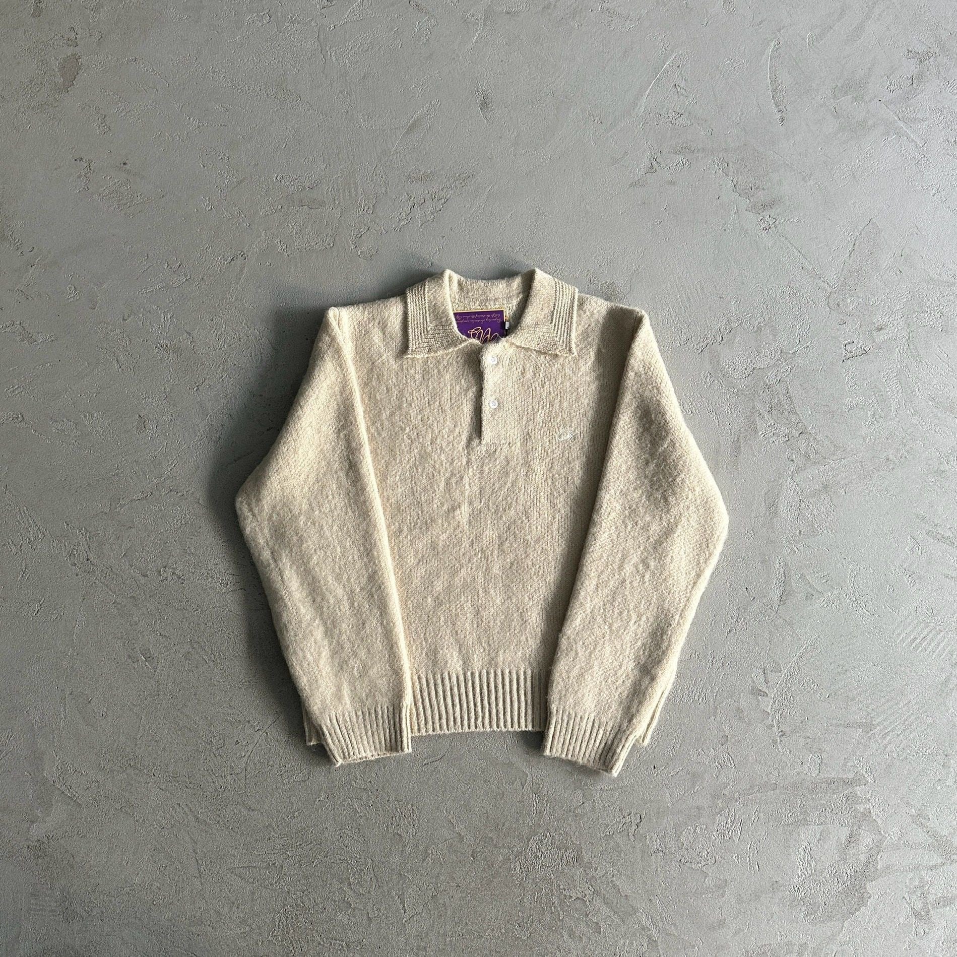 A cream-colored knit polo sweater with a short button placket, laid flat on a textured gray concrete surface.