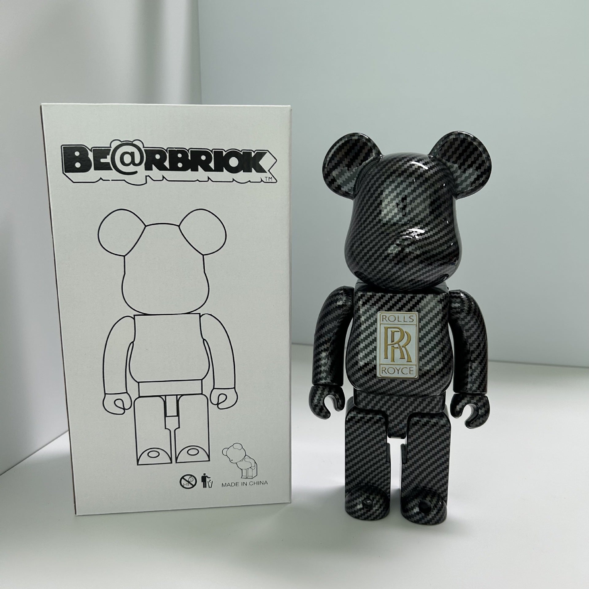 A black carbon-fiber-patterned Bearbrick figure stands next to its white packaging box. The box features the 'BE@RBRIOK' logo and a line drawing of the bear figure.