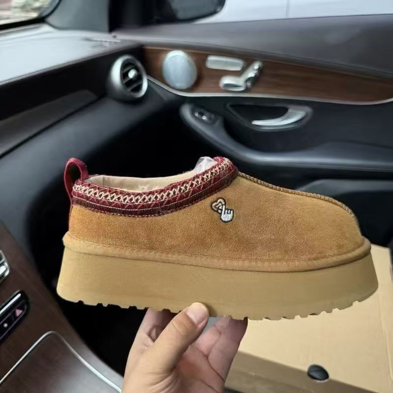 A tan suede slip-on platform sneaker with a red braided trim and a small hand-shaped metal emblem, held by a hand in front of a car interior.