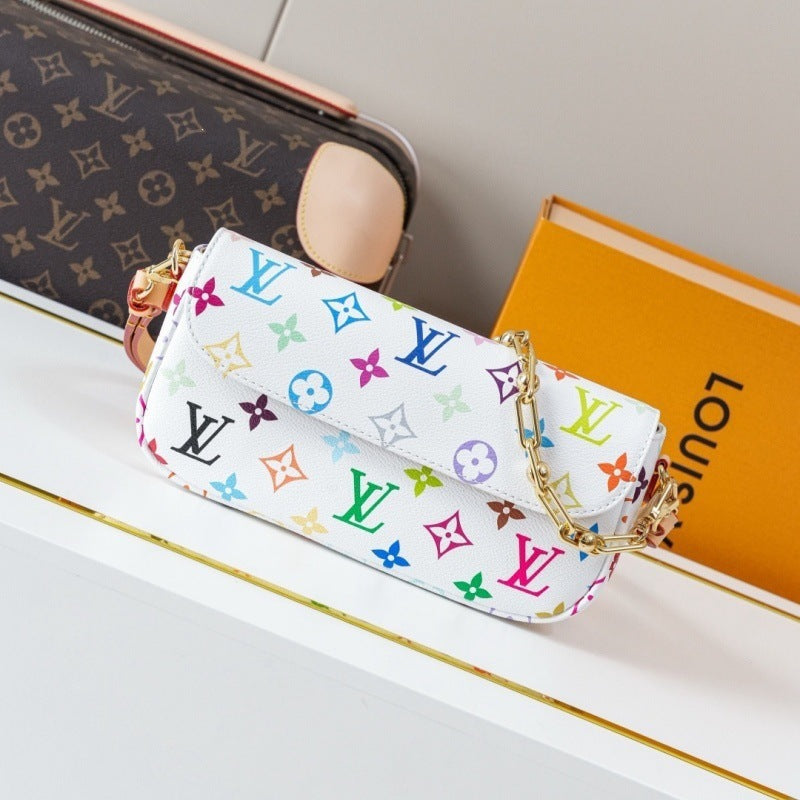 A white Louis Vuitton shoulder bag covered in a colorful monogram pattern, styled on a white shelf next to a brown Louis Vuitton monogram pouch and an orange Louis Vuitton box.