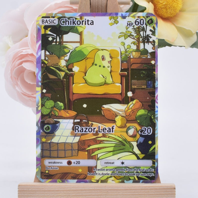 A holographic Pokémon trading card of Chikorita, displayed on a small wooden stand. The card features a colorful, illustrated interior scene with Chikorita sitting in a yellow chair, and is framed by soft pink and yellow flowers in the background.