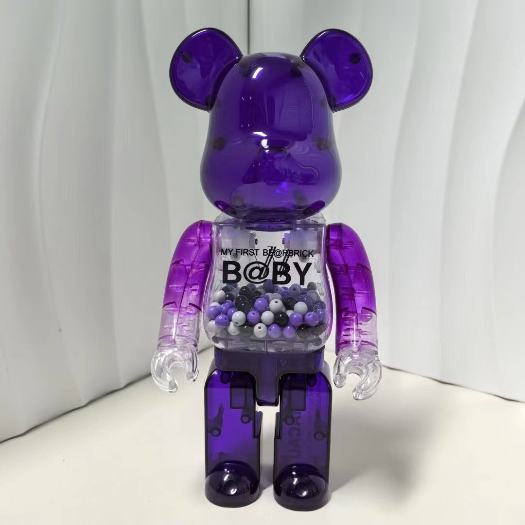 A translucent purple Bearbrick toy figure with a clear chest section filled with small beads, standing against a white background. The chest has printed text reading 'MY FIRST BEARBRICK B@BY'.