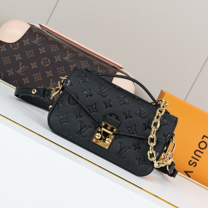A black Louis Vuitton monogram shoulder bag with gold-tone hardware, displayed on a white surface next to its orange Louis Vuitton box and a monogram pouch.