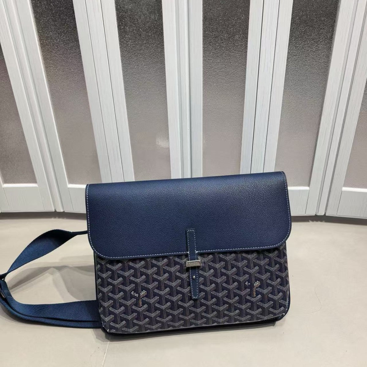 A Goyard blue crossbody bag with a navy monogram patterned body and a solid blue flap, resting on a light floor in front of white paneled doors.