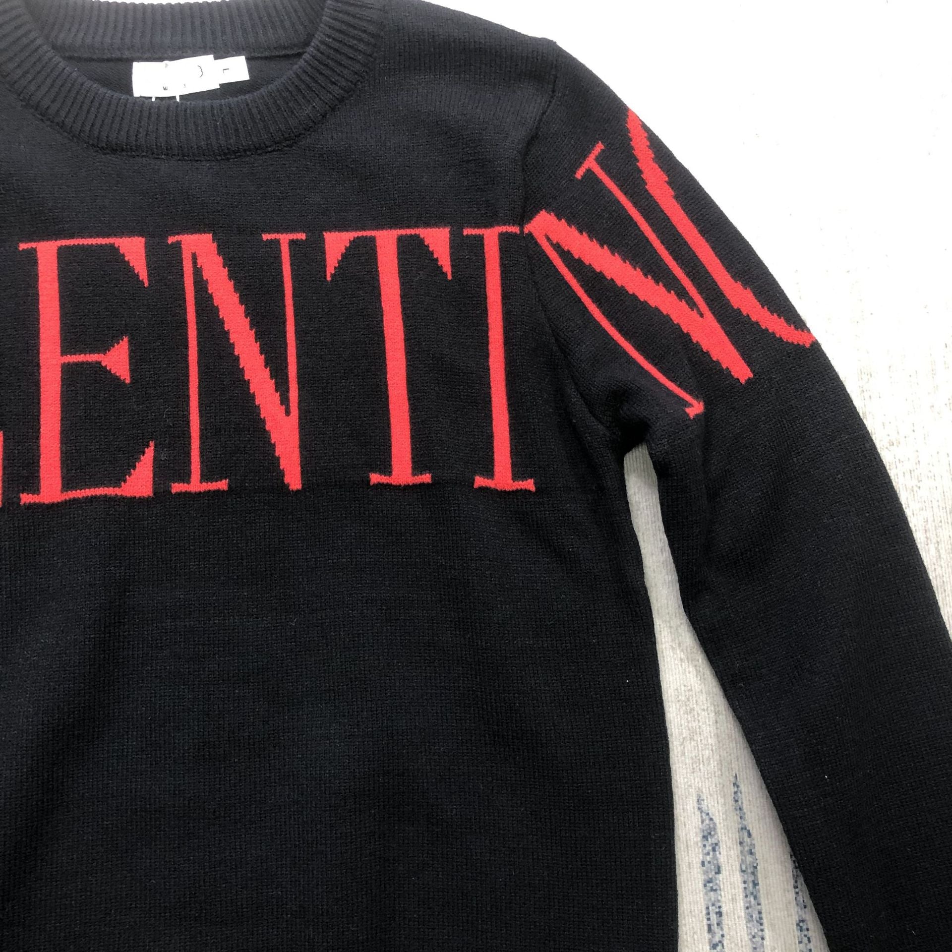 A close-up of a black knit sweater featuring a bold red knitted 'ENTIN' text across the chest and a matching red 'V' on the sleeve, with a visible Valentino neck label.