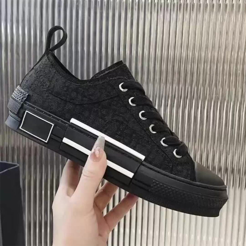 A hand holds a black low-top canvas sneaker with a textured fabric upper, black laces, a black rubber toe cap and sole, and two prominent white stripes on the side, set against a metallic silver ribbed background.