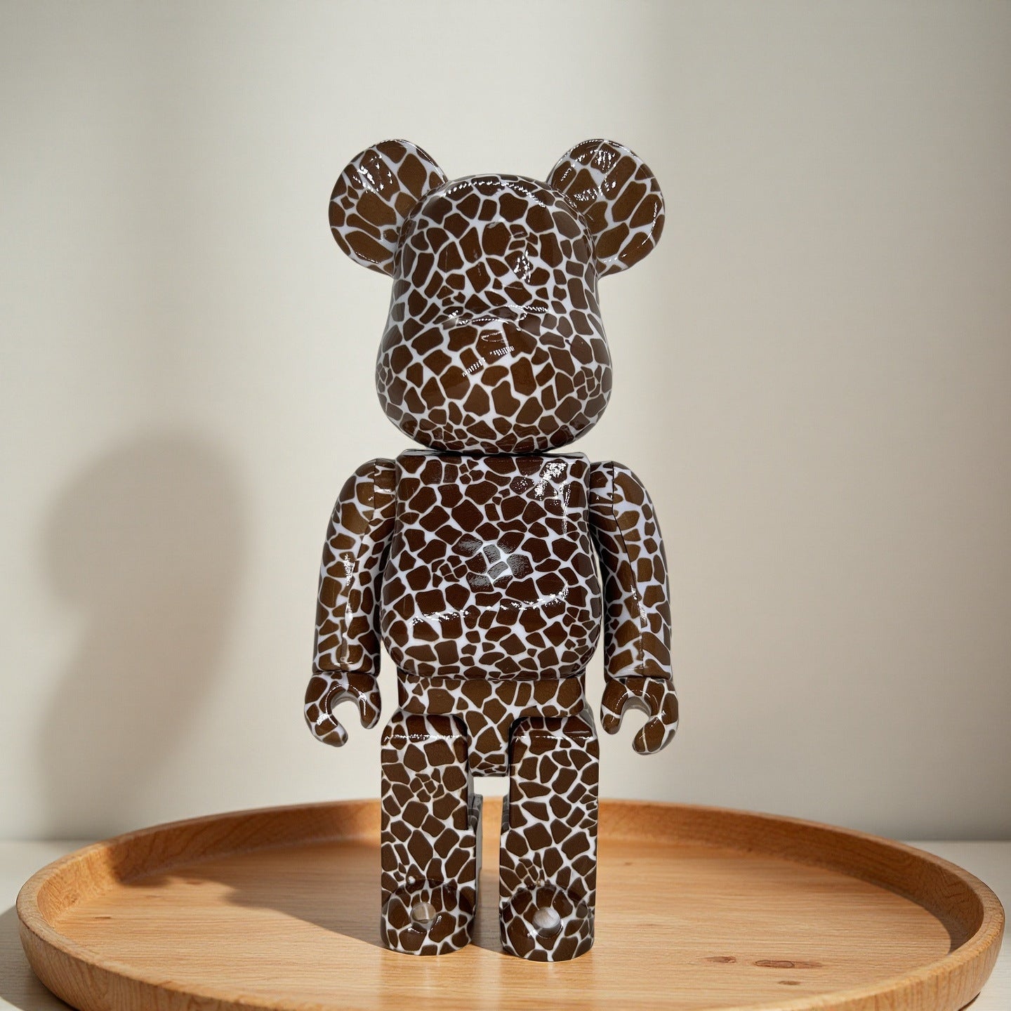 A 400% bear figure with a glossy brown and white giraffe-like spot pattern, standing on a round wooden tray against a neutral beige background.