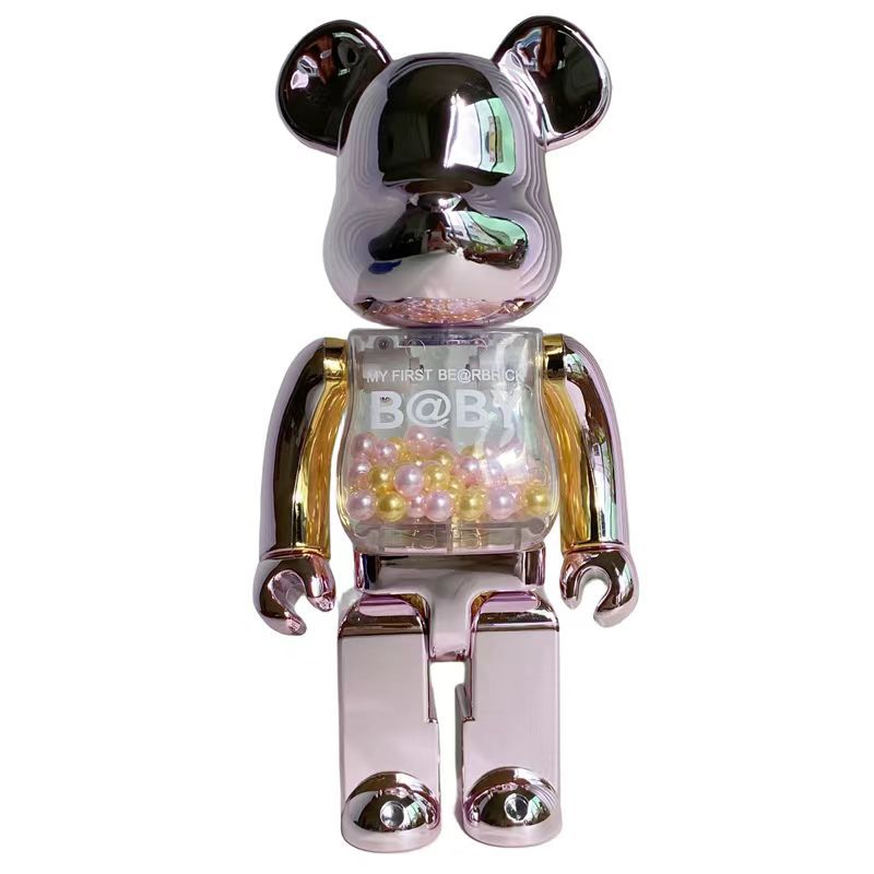 A metallic pink and gold Bearbrick toy figure with a reflective chrome finish, featuring a clear chest compartment filled with iridescent pearls. The text 'MY FIRST BEARBRICK B@BB' is printed on the chest, set against a plain white background.