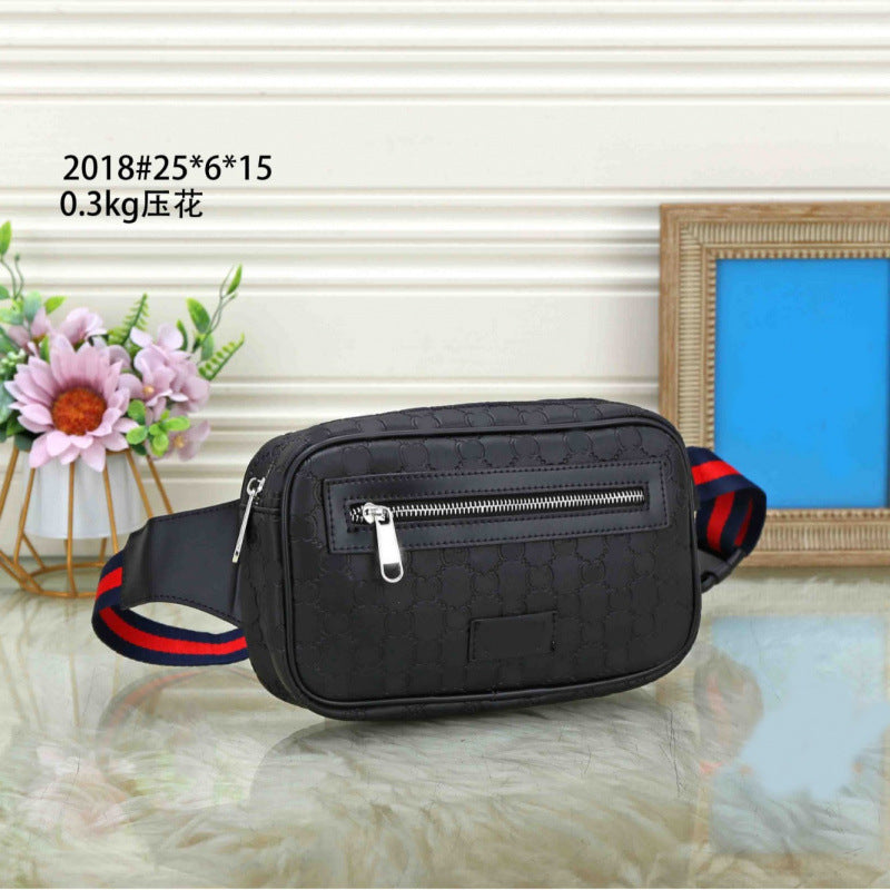 A black crossbody bag with a textured monogram pattern, featuring a front zippered pocket and a red and blue striped webbing strap. The bag is displayed on a reflective surface with decorative props, including a vase of pink flowers and a picture frame, against a cream-colored paneled wall.