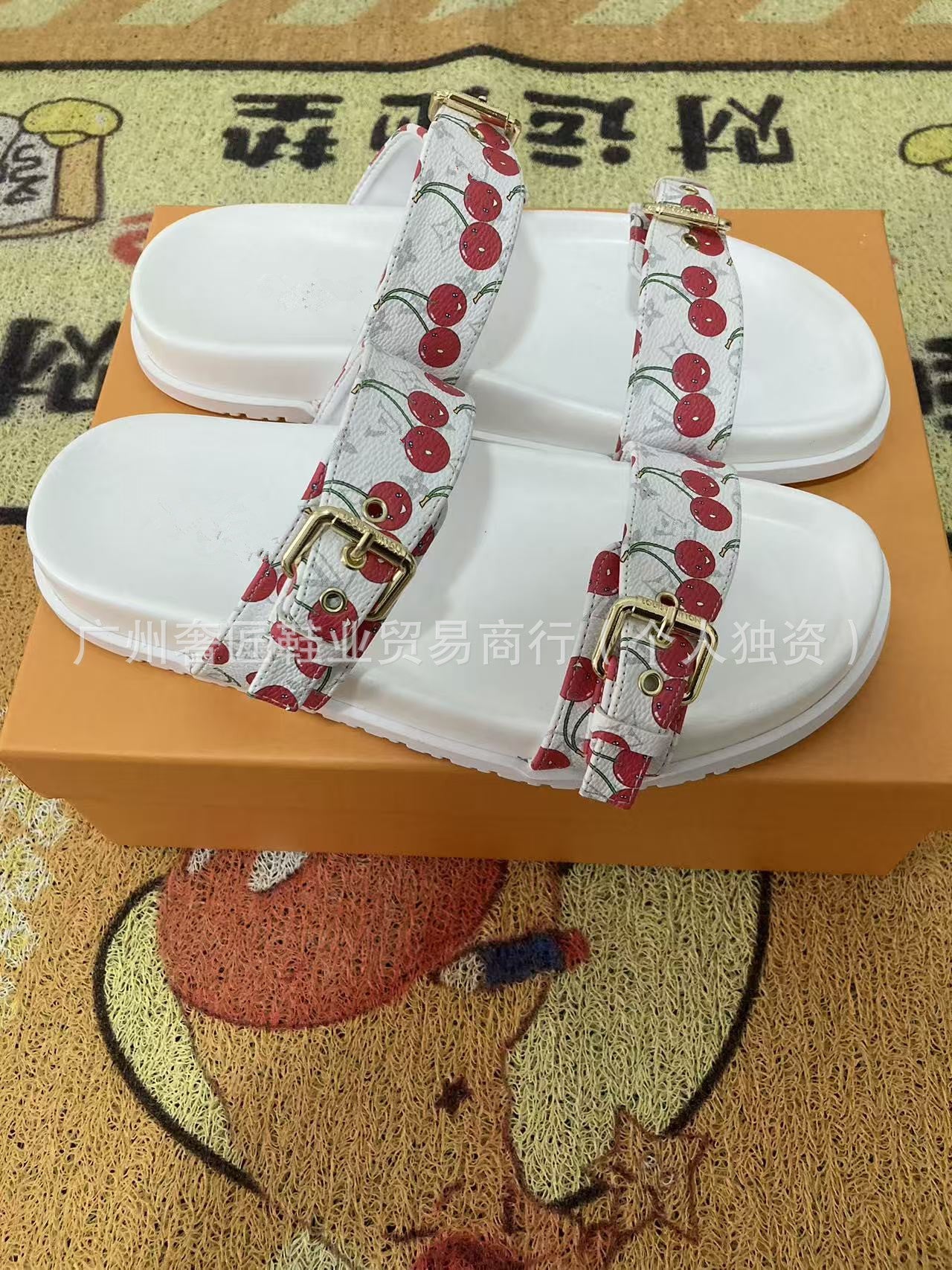 White sandals with cherry-patterned straps on an orange box, on a textured surface.