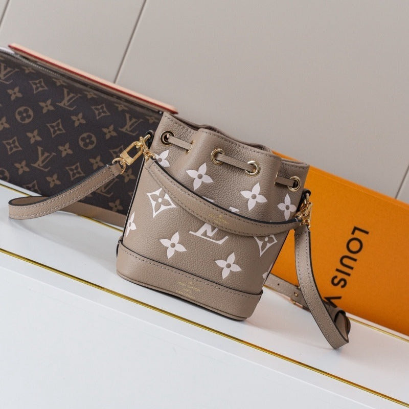 A Louis Vuitton bucket bag in taupe beige with a white monogram pattern, displayed on a white surface with its signature orange box and a dark brown monogram clutch in the background.