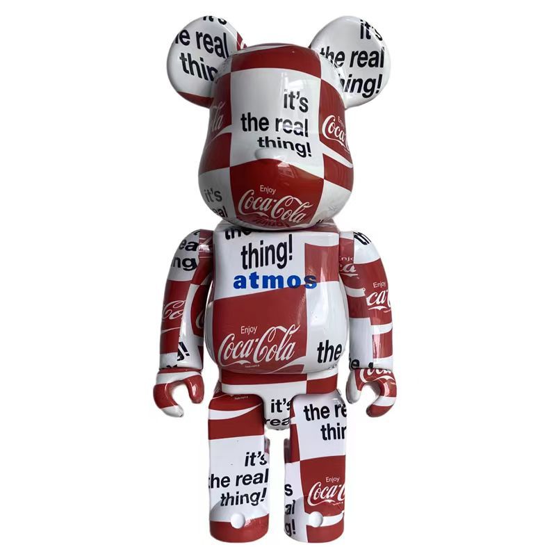A Coca-Cola branded bear-shaped collectible figure with a red and white checkerboard pattern, featuring Coca-Cola logos and phrases like 'It's the real thing!' on a plain white background.