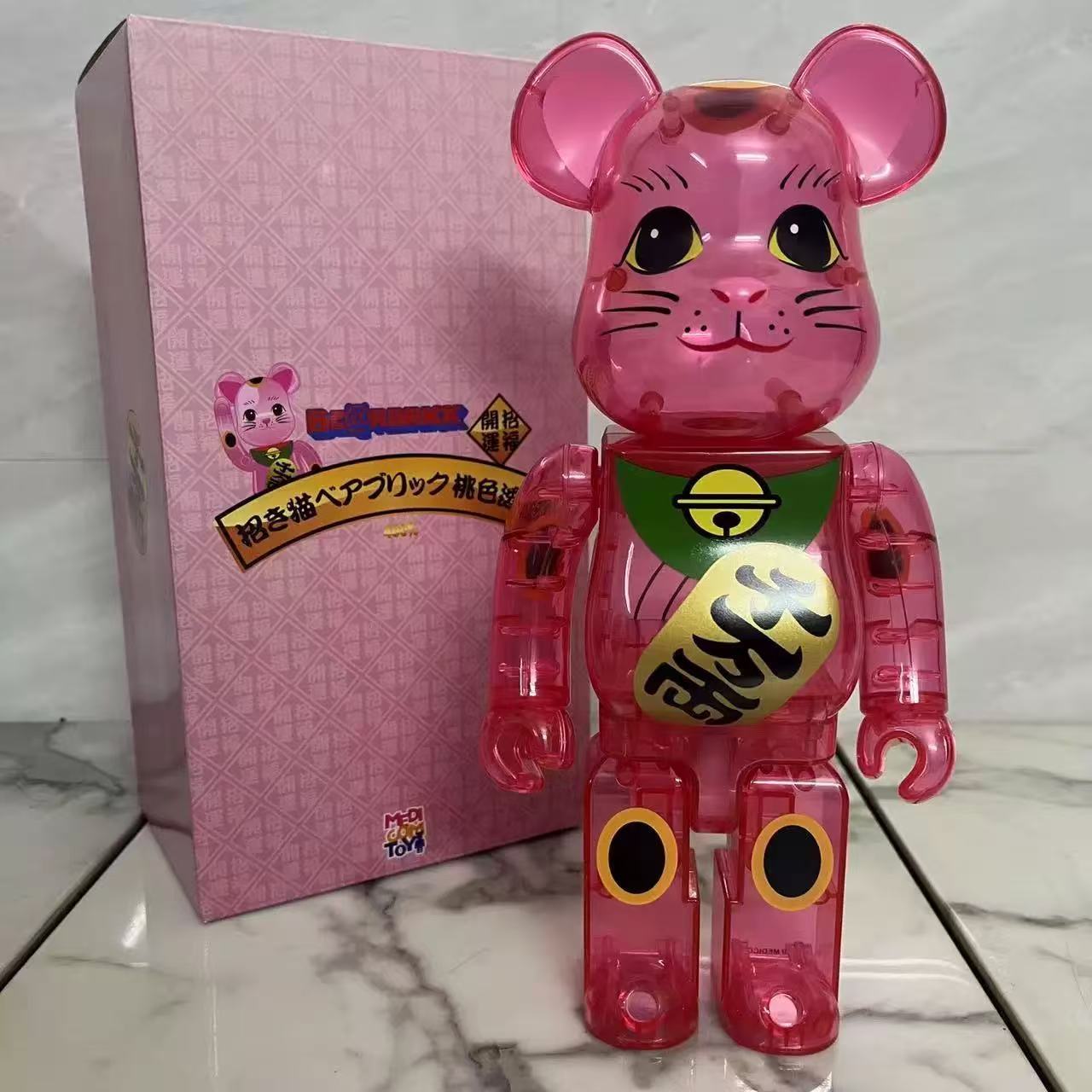 A translucent pink bear-shaped collectible figure with Japanese kanji on its chest stands in front of a pink patterned box on a white marble surface.