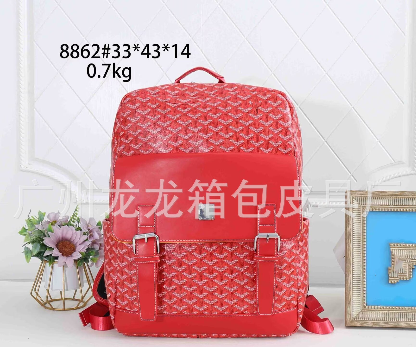 A red patterned backpack with a glossy front pocket and two buckle straps, displayed on a white surface with decorative flowers and a picture frame. Text overlays show the model number and dimensions.