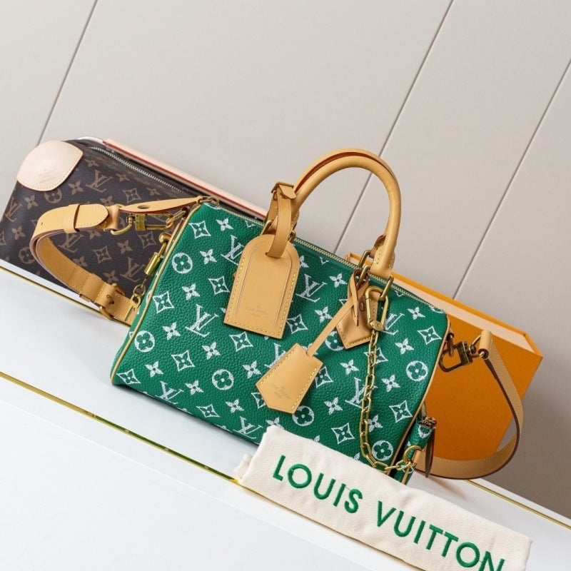 A Louis Vuitton monogram Speedy bag in green coated canvas with tan leather trim and handles, displayed on a white shelf with a Louis Vuitton dust bag in front and another bag in the background.