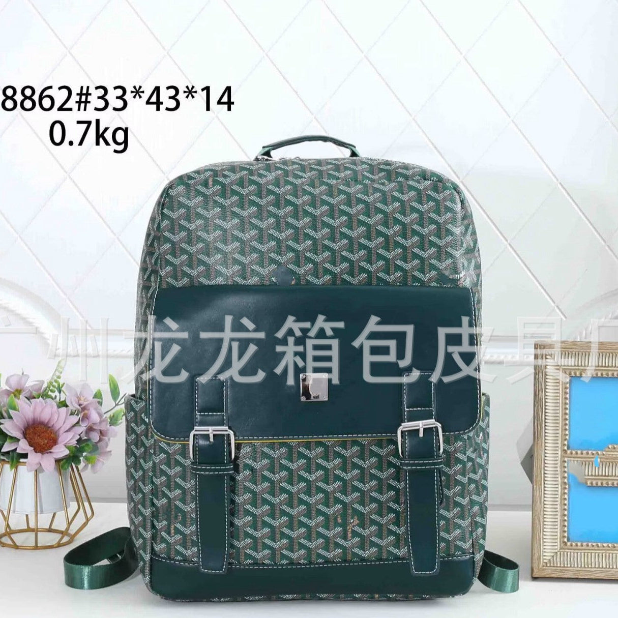 A Goyardine backpack in dark green and beige, featuring a dark green leather front pocket with two buckled straps, set against a white tiled background with decorative props.