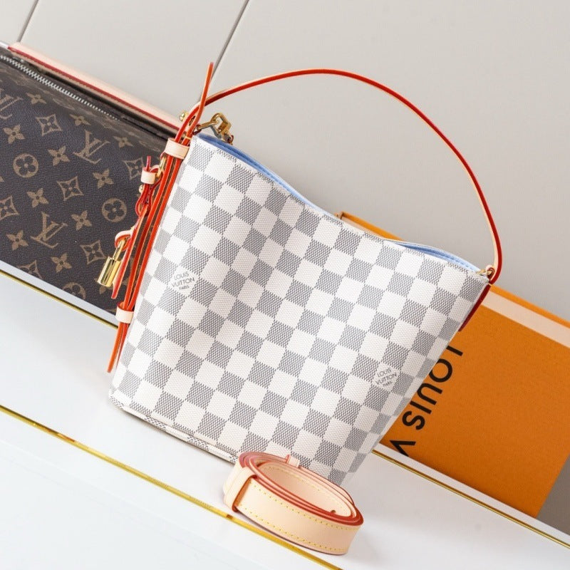 A Louis Vuitton shoulder bag in Damier Azur canvas with a white and gray checkered pattern, featuring a red leather handle and tassel, displayed with its orange box and a matching monogram pouch.