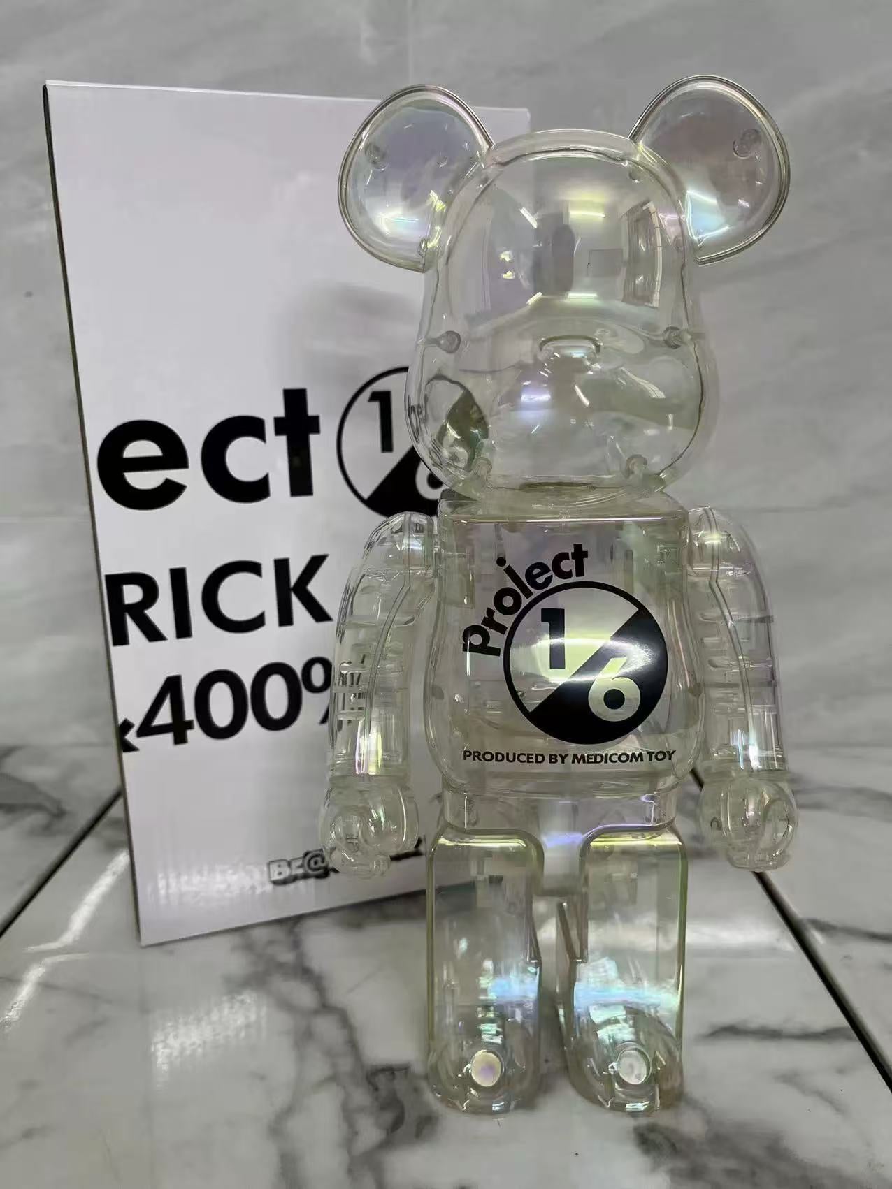 A transparent iridescent bear figure with a black and white Project Bearbrick logo on the chest, standing in front of its white packaging box on a marble surface.