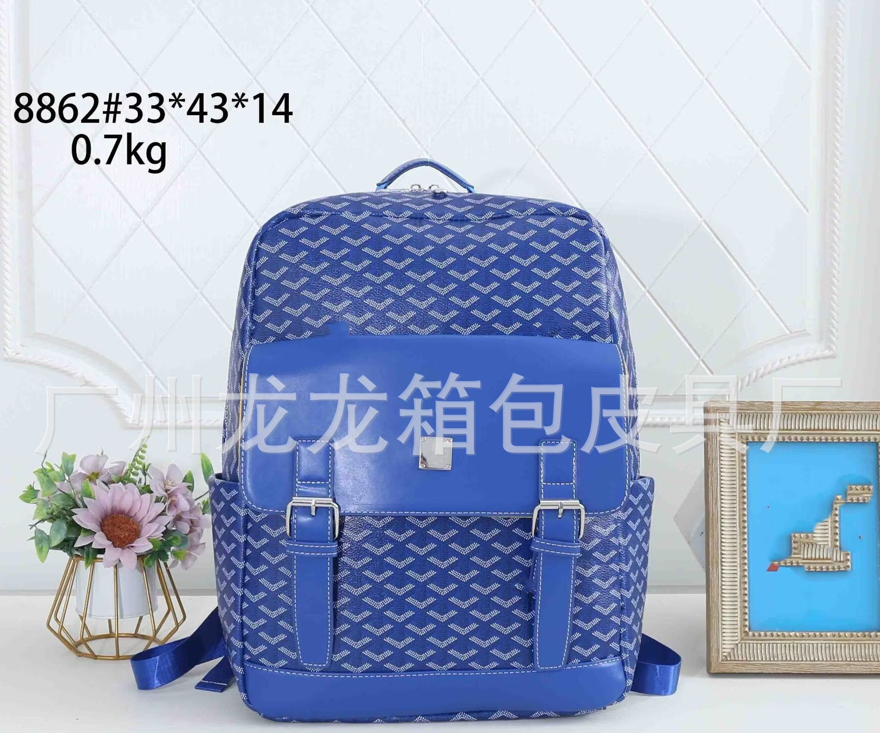 A royal blue patterned backpack with a glossy front pocket and silver buckles, displayed on a white surface with a small flower arrangement and a framed picture in the background. Text overlays show the product code and dimensions.