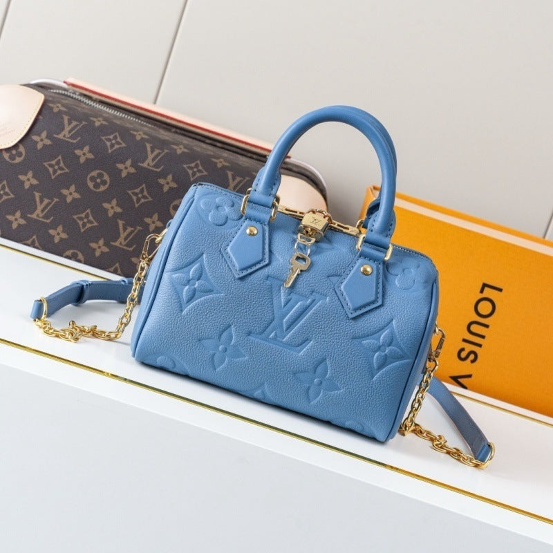 A light blue Louis Vuitton handbag with embossed monogram pattern and gold-tone hardware, styled on a white surface with a Louis Vuitton box and a monogram pouch in the background.