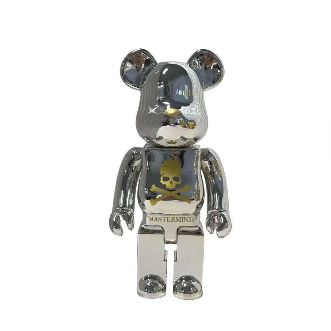 A metallic silver Bearbrick figure with a reflective chrome finish, featuring a gold skull and crossbones graphic on the chest and the brand name 'MASTERMIND' printed below it, set against a plain white background.
