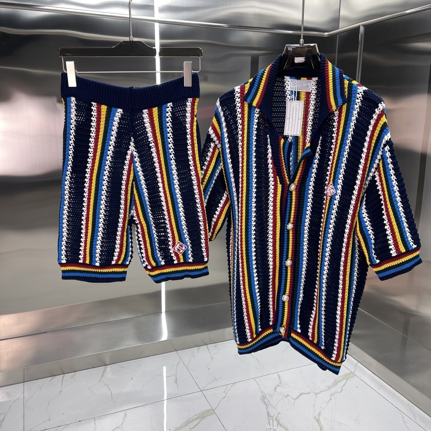 Colorful knitted cardigan and shorts set on hangers against a reflective floor and wall.