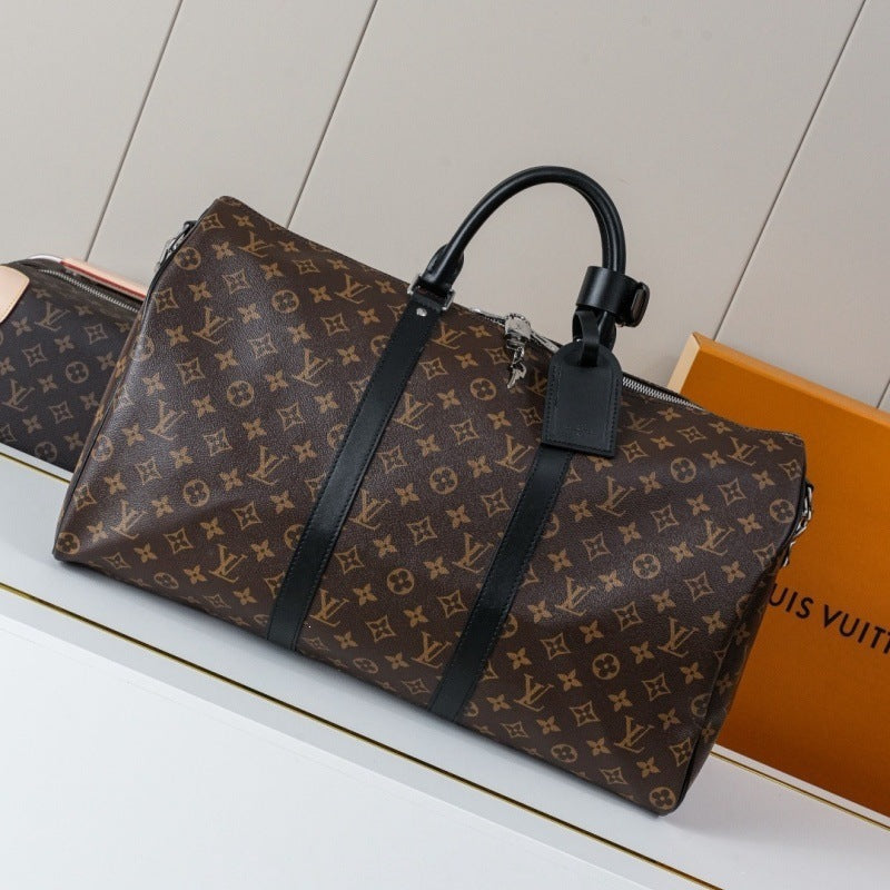 A Louis Vuitton monogram duffle bag in brown coated canvas with black leather handles and trim, displayed on a white surface next to an orange Louis Vuitton box.