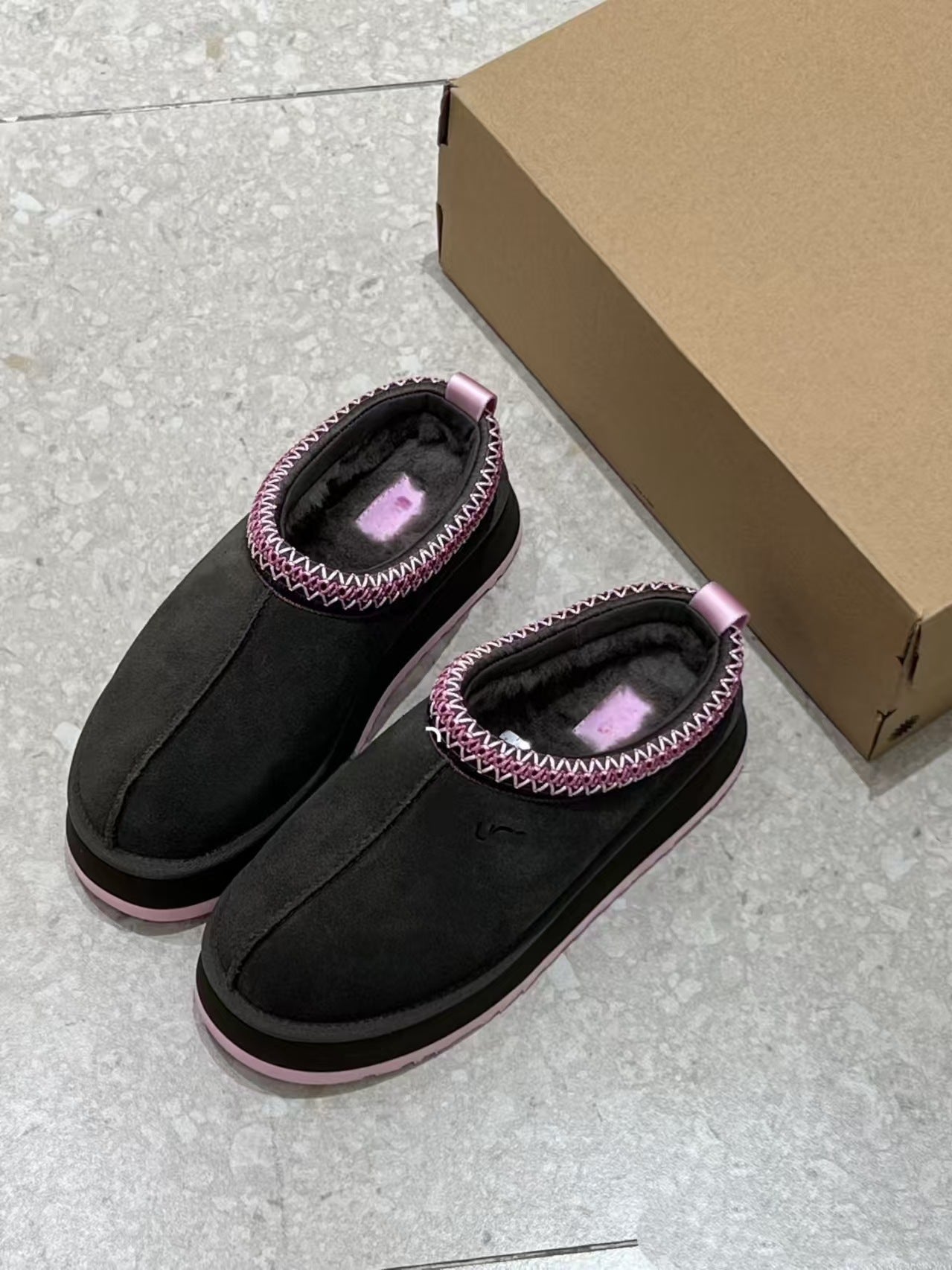 A pair of black slip-on slippers with pink and white zigzag trim and pink soles, shown next to a brown cardboard box on a light gray tiled floor.