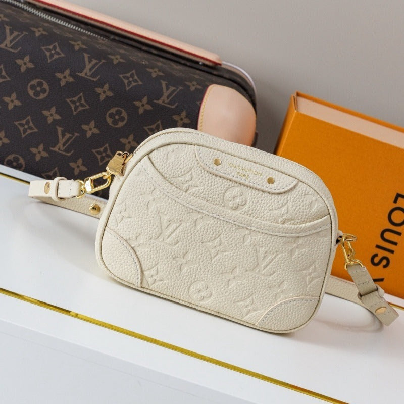 A Louis Vuitton monogram crossbody bag in cream-colored pebbled leather with gold-tone hardware, displayed on a white surface next to its orange Louis Vuitton box and a brown monogram pouch.