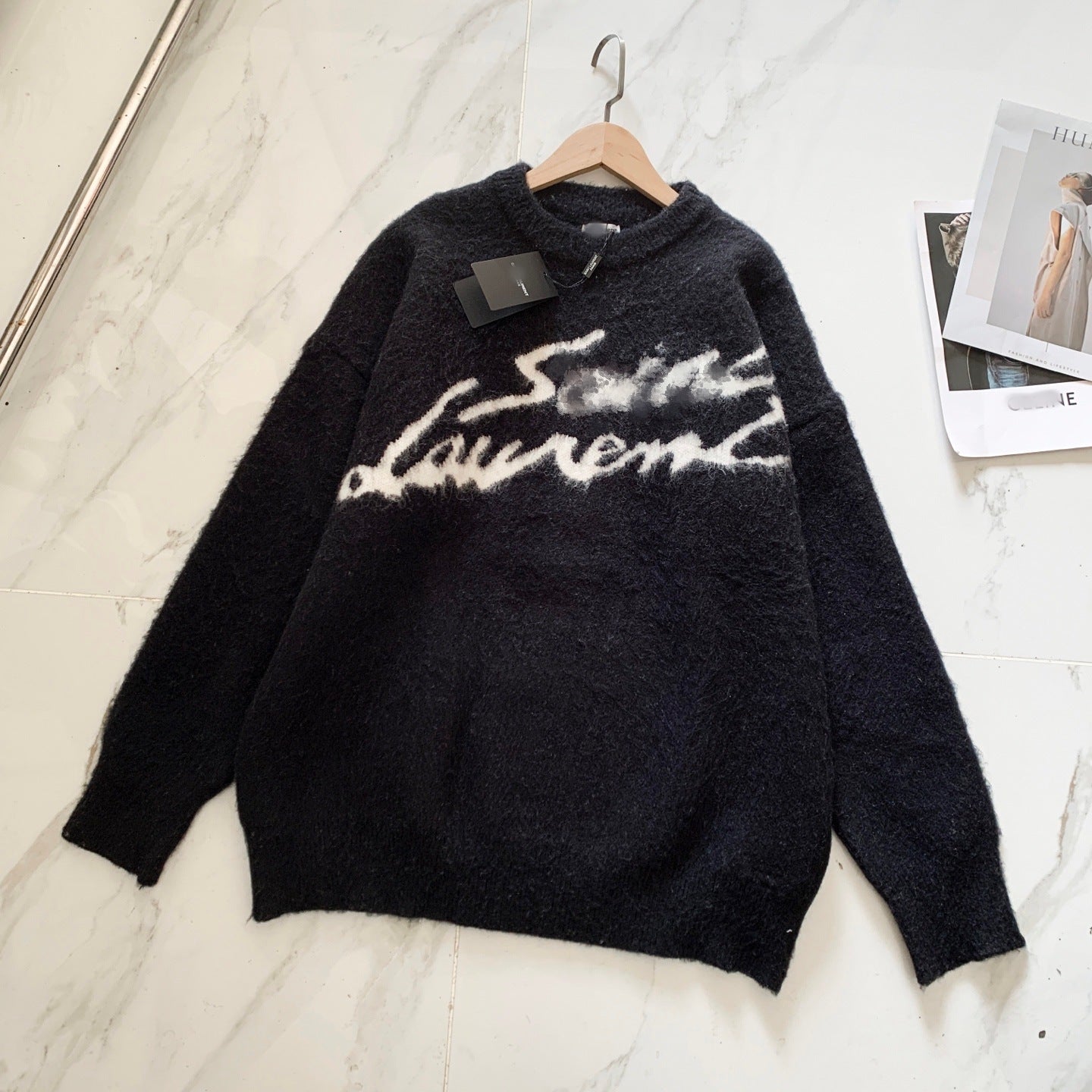 A black fuzzy knit sweater with a white 'Saint Laurent' script across the chest, hanging on a wooden hanger against a white marble-patterned background.